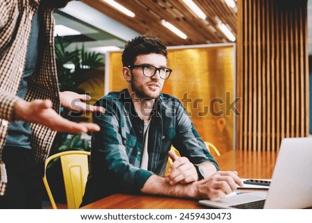 Cropped image of young bearded surprised graphic designer and pondering colleague discussing ideas spending time in coworking space.Talented hipster guys collaborating together in office using laptop