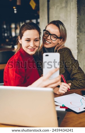 Happy female bloggers making selfie photos on modern smartphone during teamworking on successful project at laptops.Positive hipster girls taking pictures on front camera of cellular with 4G internet