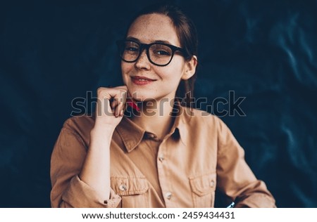 Half length portrait of happy female graphic designer in eyeglasses smiling at camera while holding smartphone in hands and working remotely at digital laptop computer connected to wireless internet