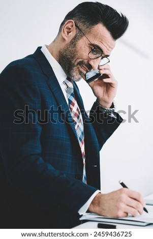 Successful mature businessman in formal wear signing financial documents of accounting reports while laughing during phone conversation on smartphone.Positive proud ceo middle aged calling on cellular