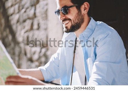Cropped image of cheerful bearded man traveler in stylish black sunglasses laughing while searching destination of showplaces and route on map sitting outdoors on street of old architectural city