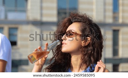 Close up of beautiful young Caucasian woman with curly hair in sunglasses drinking beer on sunny day outdoor at a party. Happy pretty stylish female with drink dancing in good mood