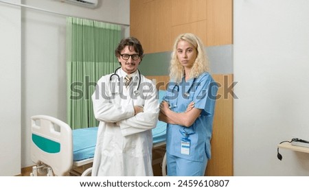 Portrait of a confident doctor in white gown and stethoscope standing with arms crossed in a hospital patient room with young nurse in blue uniform