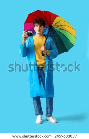 Young man in raincoat with rainbow umbrella and photo camera on blue background