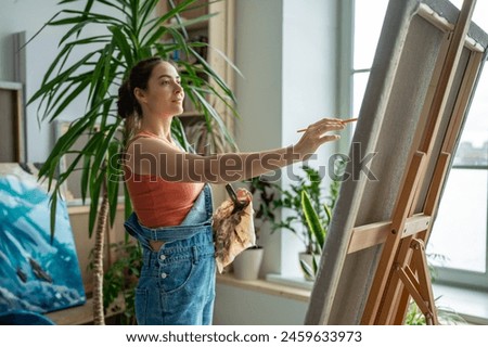 Happy smiling artistic female working on painting. Outstretched hand of satisfied glad woman with brush reaches for canvas, making careful thoughtful brushstrokes, creating masterpieces, artworks.