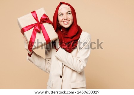 Young Arabian Asian Muslim woman wear red abaya hijab suit clothes hold present box with gift ribbon bow look aside isolated on plain beige background studio UAE middle eastern Islam religious concept