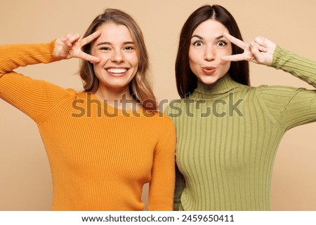 Close up young happy friends two women they wear orange green shirt casual clothes together cover eyes with v-sign look camera isolated on plain pastel light beige background studio. Lifestyle concept