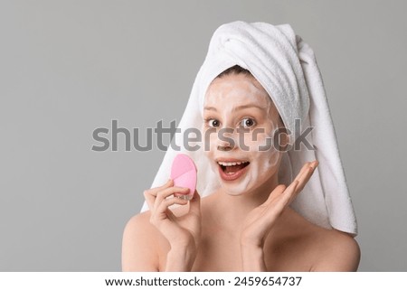 Emotional young woman washing face with brush and cleansing foam on grey background, space for text