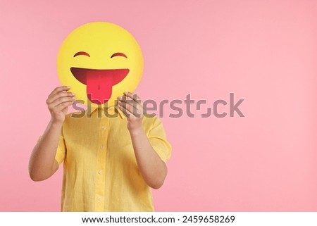 Woman covering face with emoticon sticking out tongue on pink background. Space for text
