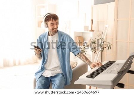 Young tattooed man in headphones with mobile phone playing synthesizer at home Royalty-Free Stock Photo #2459664697