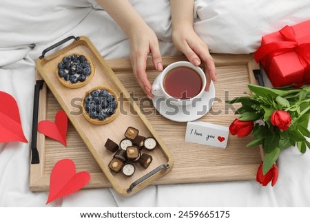 Tasty breakfast served in bed. Woman with tea, desserts, gift box, flowers and I Love You card at home, above view