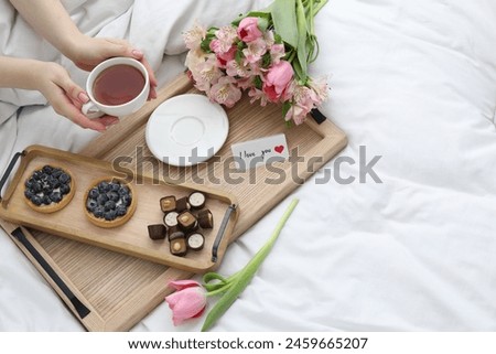 Tasty breakfast served in bed. Woman with tea, desserts, flowers and I Love You card at home, above view. Space for text