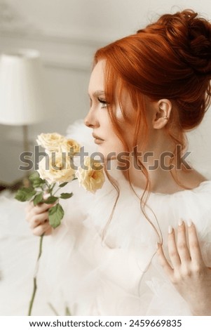 Elegant lady with bright red hair, clad in a white dress, posing gracefully with a bouquet of white roses indoors