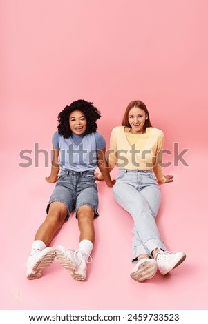 Two diverse women in casual attire sitting on a pink background, posing for a picture.