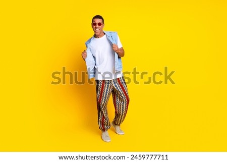 Full length photo of good mood stylish guy in sunglass dressed print pants holding jeans waistcoat isolated on yellow color background