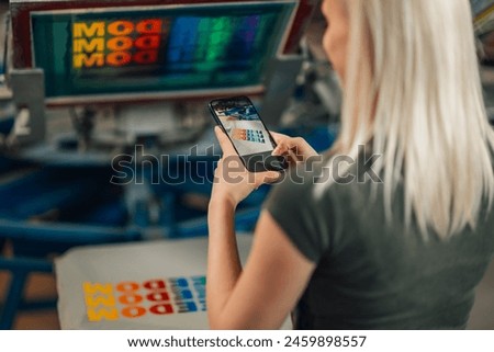 Cropped picture of female entrepreneur taking phone photos of silkscreen printed t-shirt in printing workshop. Close up of female graphic technician taking pictures of screen printed textile product.