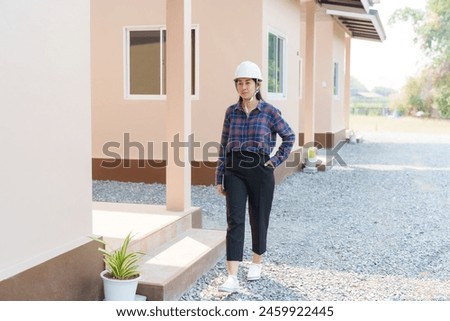 Asian woman in plaid shirt, hard hat, stands confidently outside house, hands on hips, ready to inspect.Inspectors inspect the completed house.


