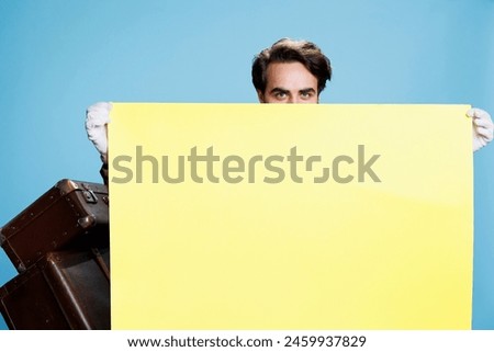 Doorkeeper presents empty ad banner in studio, young man works as hotel porter. Classy elegant bellboy showing blank yellow poster to do advertisement sign on camera, travel industry.