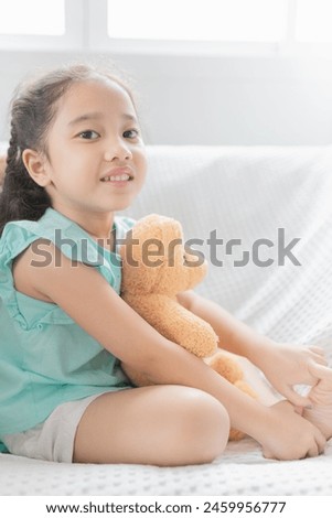 attractive asian children sitting on white sofa, she hug bear doll and feeling happy in the morning time