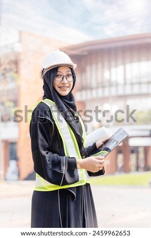 A beautiful Asian Muslim construction engineer portrait in safety vest and Abaya niqab traditional clothes standing and hold a tablet computer on modern building background.