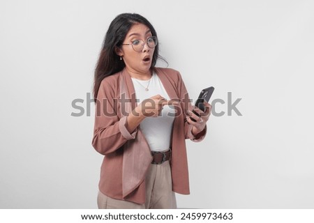 Shocked Asian business woman in eyeglasses pointing to her smartphone, isolated by white background.