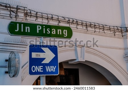 An image capturing a vibrant Jln Pisang street sign next to a blue one way directional arrow on a white architectural background with security features. Translated: the banana street. Royalty-Free Stock Photo #2460011583