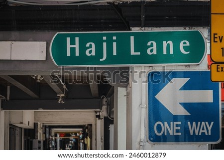 A vivid image displaying a green street sign for Haji Lane next to a blue one-way directional arrow under an urban bridge.