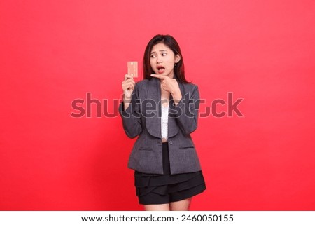 Asian office woman's expression shocked candid hand holding a debit credit card while pointing at it wearing a jacket and skirt on a red background. for financial, business and advertising concepts