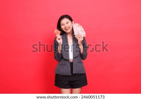 gesture of cheerful Asian office woman holding credit card and money in head area wearing jacket and skirt on red background. for transaction, business and advertising concepts