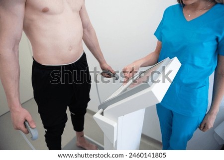 Shirtless man stands beside a medical device as a nurse assists in assessing his body metrics