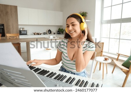 Beautiful young happy woman in headphones listening music and playing synthesizer at kitchen Royalty-Free Stock Photo #2460177077