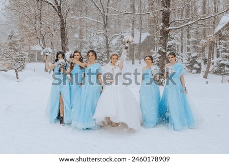 A bride and her bridesmaids are posing for a picture in the snow. The bride is wearing a white dress and the bridesmaids are wearing blue dresses. Scene is joyful and festive, as the bride