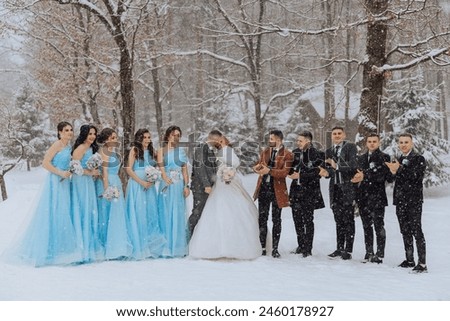 A bride and groom are surrounded by their wedding party in the snow. The bride is wearing a blue dress and the groom is wearing a brown coat. The wedding party is posing for a picture