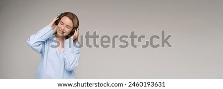 Enjoying the music with eyes closed and wide smile.  blonde woman in blue shirt touching her black headphones on her neck.   long horizontal banner Gray background studio shot 