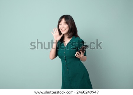 An Asian woman green dress shouting gesture to free copy space while holding mobile phone isolated on pastel green background. online payment, promotion, deals concept.