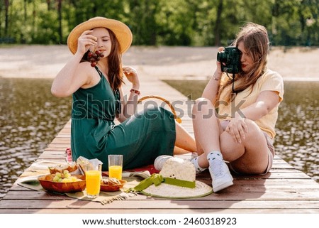 A woman in a yellow sun hat is happily taking a picture of another woman having a picnic on the grass with tableware under the bright sun, using flash photography