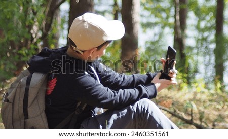 teenage boy with a backpack in the forest, taking a selfie on a smartphone, shooting a video for a blog, content. catching signal at wooden forest. Road trip, travel, technology internet and people.