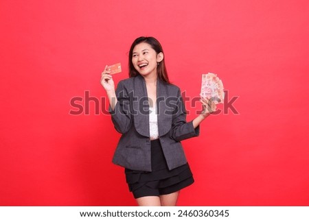 gesture of happy candid asian office girl holding credit card in front and money wearing jacket and skirt on red background. for financial, business and advertising concepts