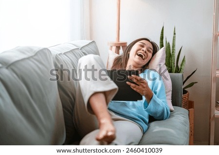 enjoyment, lying, cute, rest, living, cozy, pretty, relaxing, couch, comfortable. A woman is sitting on a couch with a tablet in her lap. She is smiling and laughing while using the tablet. Royalty-Free Stock Photo #2460410039