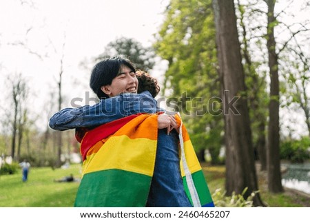 A Chinese man and his Latino partner share a heartwarming hug, enveloped in a colorful LGBT flag Royalty-Free Stock Photo #2460465207