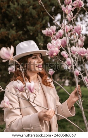 Woman magnolia flowers, surrounded by blossoming trees., hair down, white hat, wearing a light coat. Captured during spring, showcasing natural beauty and seasonal change.