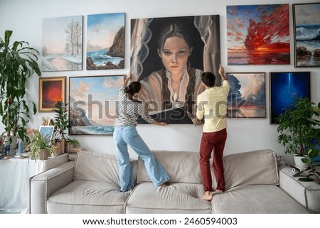 Artists couple hang picture on wall. Man, woman standing on sofa couch creating cozy creative inspiring environment atmosphere at home art studio. Spouses posting painting artwork at wall together