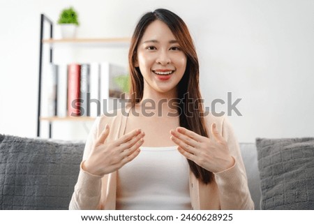 A young deaf woman is sitting on a couch and making a sign Language to communicate. She is smiling and she is happy