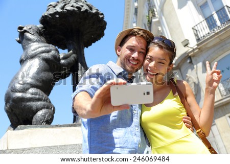 Tourists taking selfie photo pictures by famous bear statue Madrid on Puerta del Sol. Young couple using smartphone camera at tourist attraction Bear and the Madrono Tree, symbol of Madrid, Spain.