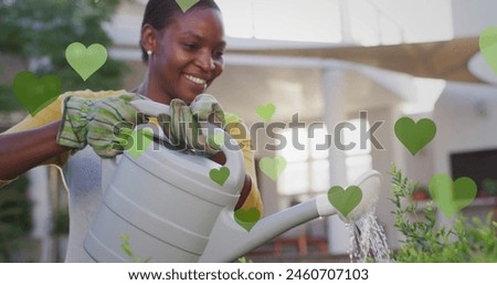 Image of green hearts over happy african american woman watering plants. houseplants, taking care of plants and hobby concept digitally generated image.
