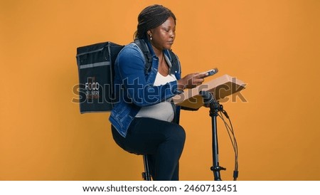 Black woman delivers takeout orders on bicycle, utilizing wireless payment device for seamless transactions. African american delivery person taking pos machine from pocket for contactless pay. Royalty-Free Stock Photo #2460713451