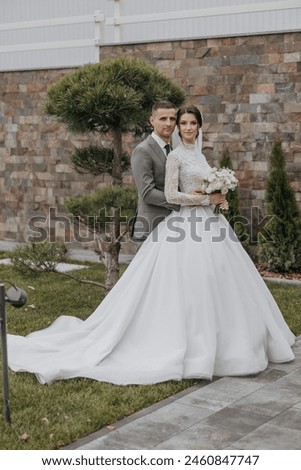 A bride and groom are posing for a picture in front of a tree. The bride is holding a bouquet and the groom is holding a white flower. Scene is happy and romantic