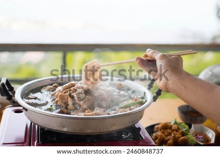 Slice of pork was picking up by the chopstick in the woman's hand above the curved brass pan around with diffused smoke. Royalty-Free Stock Photo #2460848737