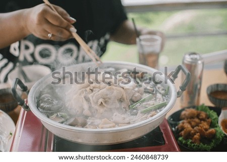 Slice of pork was picking up by the chopstick in the woman's hand above the curved brass pan around with diffused smoke. Royalty-Free Stock Photo #2460848739