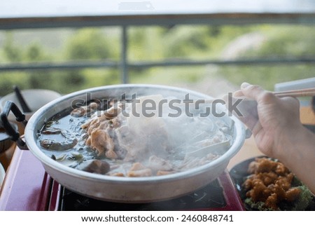 Slice of pork was picking up by the chopstick in the woman's hand above the curved brass pan around with diffused smoke. Royalty-Free Stock Photo #2460848741